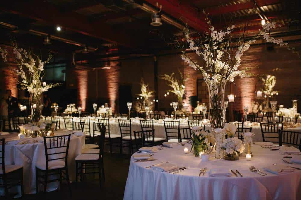 Dimly lit event space decorated for a formal dinner, featuring round tables with white tablecloths, floral centerpieces, and candles.