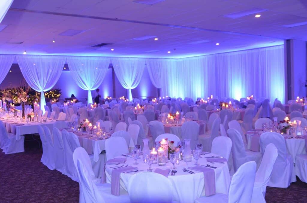 A large banquet hall illuminated in blue is decorated with white drapes, chair covers, candles, and floral centerpieces on tables.